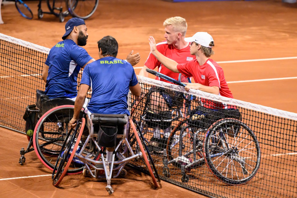 Two doubles teams in wheelchair tennis shake hands at the center of te court. 