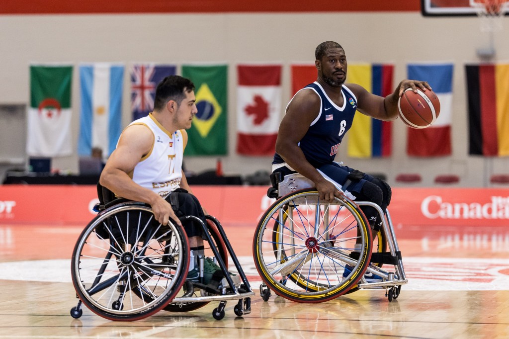 Two male wheelchair basketball players on the court, one dribbling a basketball while the other is positioned nearby, with international flags in the background.