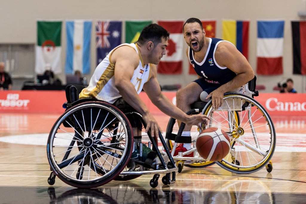 Two male athletes in wheelchairs competing in a basketball game, one reaching for the ball while the other celebrates with a loud expression. Background shows various international flags.