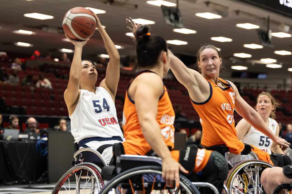 A female athlete in a wheelchair wearing a USA jersey is shooting a basketball during a competitive game, while two other players in orange jerseys are defending.
