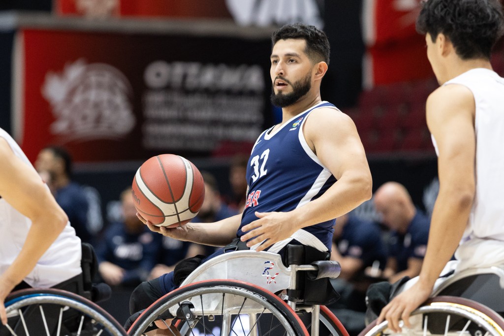 A male wheelchair basketball player in a blue uniform with 'USA' on it, holding a basketball while seated in his wheelchair, focused on the game.