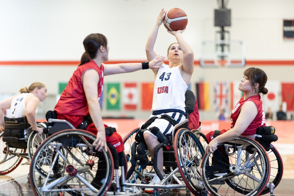 A female athlete in a wheelchair basketball game is taking a shot while two opponents attempt to block her. The player wears a white jersey adorned with 'USA' and the number 43. Flags from various countries are visible in the background.