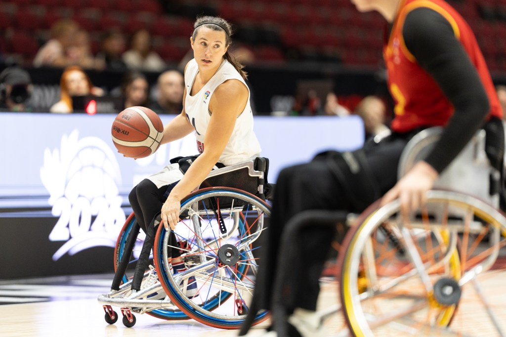 A female wheelchair basketball player dribbles a basketball while looking towards a teammate during a game.