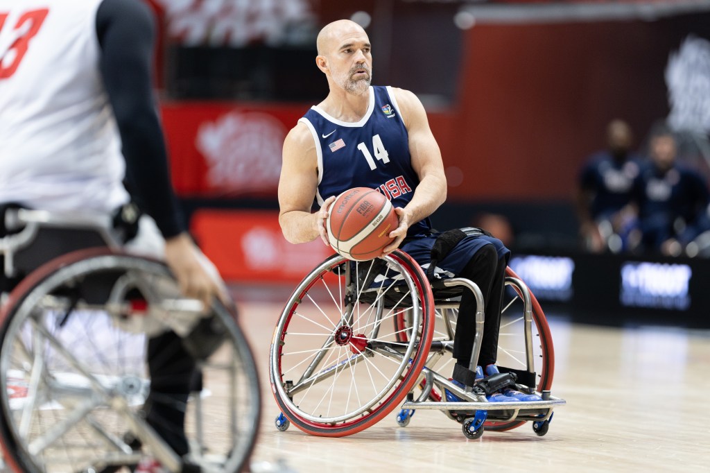 A male athlete in a wheelchair prepares to shoot a basketball during a game, wearing a navy blue jersey with the number 14 and the USA emblem.