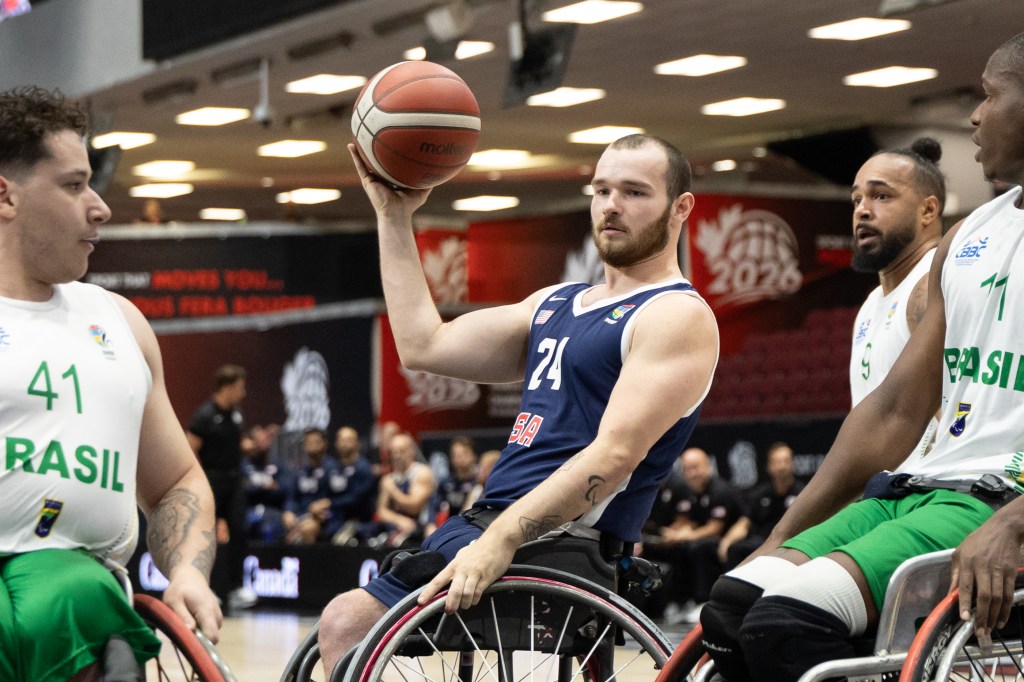 A male athlete in a wheelchair basketball game is about to shoot a basketball, while two other players are positioned nearby. One player wears a white jersey with 'BRAZIL' printed, and the other wears a dark blue jersey representing the USA.