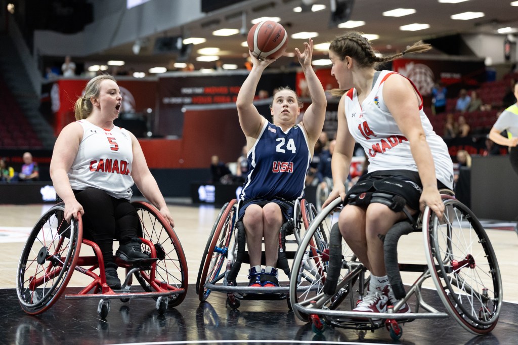 A wheelchair basketball game featuring three female athletes, two representing Canada and one representing the USA. The athlete in blue is shooting for the basket while the Canadian players are positioned nearby, one displaying a focused expression and the other cheering.