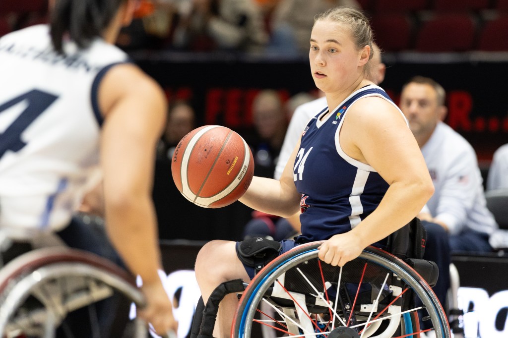 A female wheelchair basketball player in a navy uniform holding a basketball, looking determined during a game.