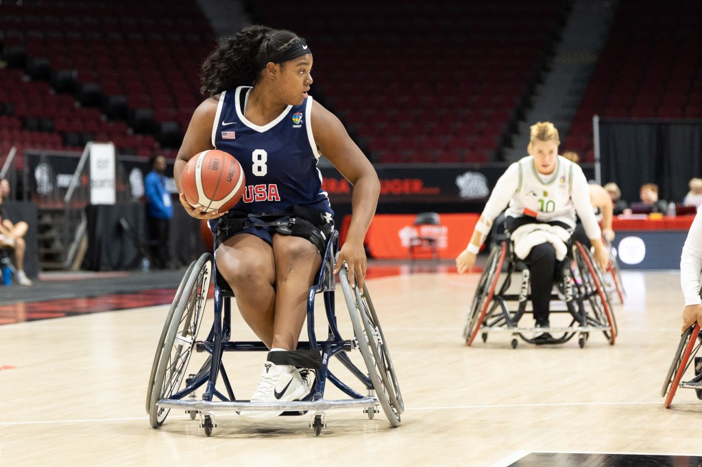 A female athlete in a wheelchair basketball game, wearing a blue jersey with 'USA' and the number 8, dribbles a basketball on the court while other players in wheelchairs are positioned nearby.