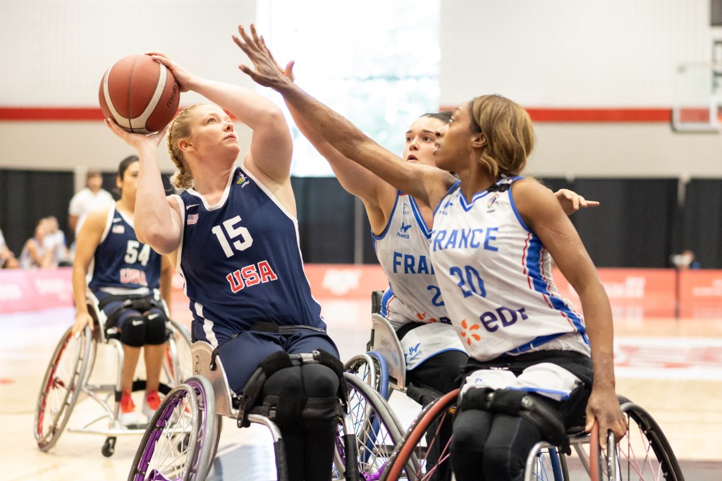 A female wheelchair basketball player from the USA, wearing jersey number 15, attempts a shot while being defended by two players from France, one wearing jersey number 20, in a competitive game.