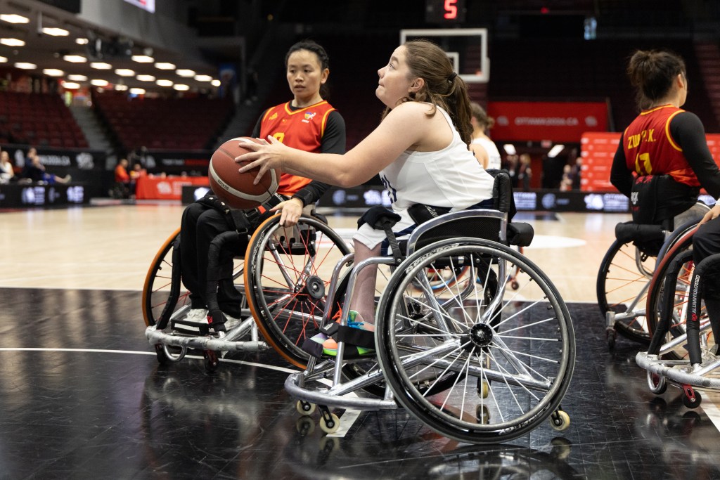 A young female athlete in a wheelchair prepares to pass a basketball during a wheelchair basketball game, while another player looks on in the background.