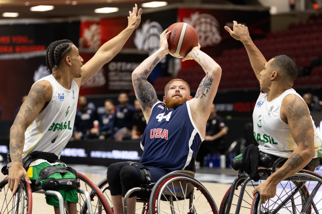 A wheelchair basketball player from the USA attempting a shot while being guarded by two Brazilian players.