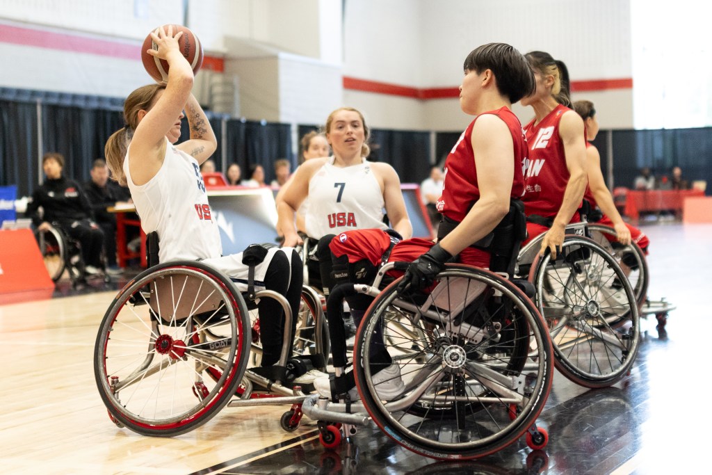 A group of female wheelchair basketball players in action, with one player holding a basketball above her head. The player in white represents the USA, while others in red represent a different team. The scene captures a moment during a competitive game.